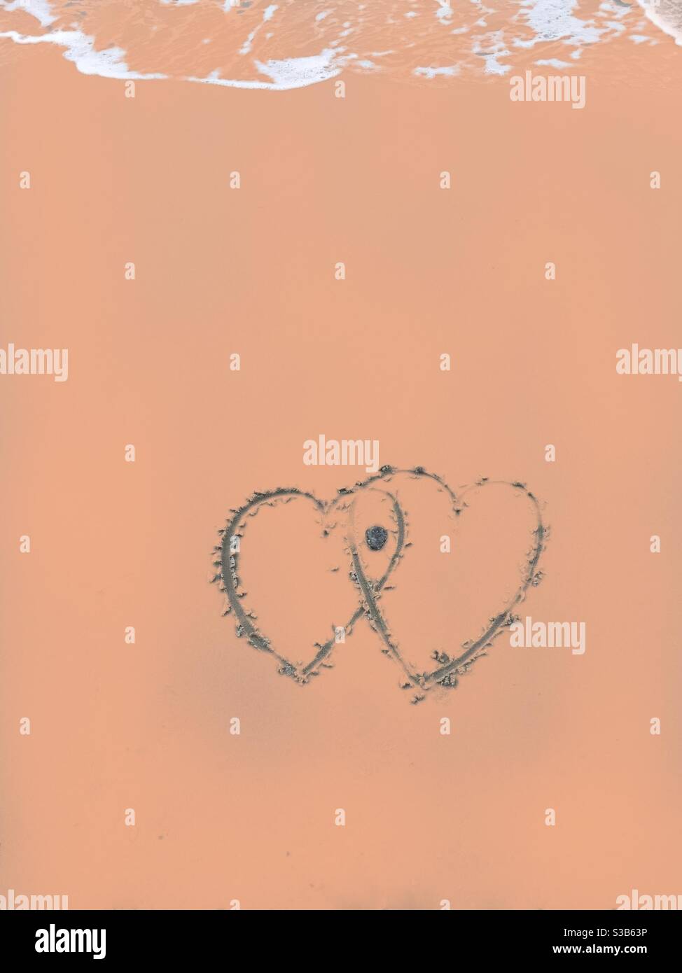 Sich schneidende Herzen, die in Sand gezogen sind, nähern sich einer Welle, die die Zeichnung bedeckt, schwächt und dann wegspült, wobei vielleicht nur der Stein ein symbolisches flinty Herz hinterlässt. - Smartphone-aufgenommenes Stockfoto