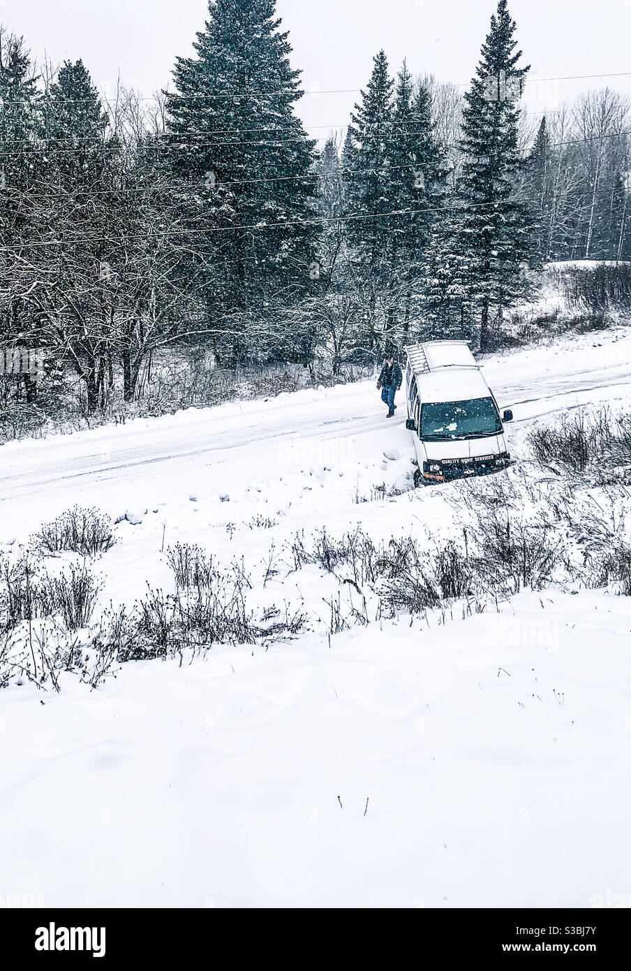 An einem verschneiten Tag steckte ein Arbeitswagen in einem Graben fest. Die Ansicht ist aus einem Fenster im zweiten Stock. Der Arbeiter läuft um seinen LKW herum und überwacht den Schaden. - Smartphone-aufgenommenes Stockfoto