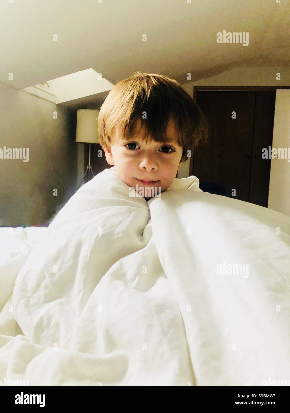 Bezauberndes Porträt eines dreijährigen Jungen in einer weißen Decke gewickelt. - Smartphone-aufgenommenes Stockfoto
