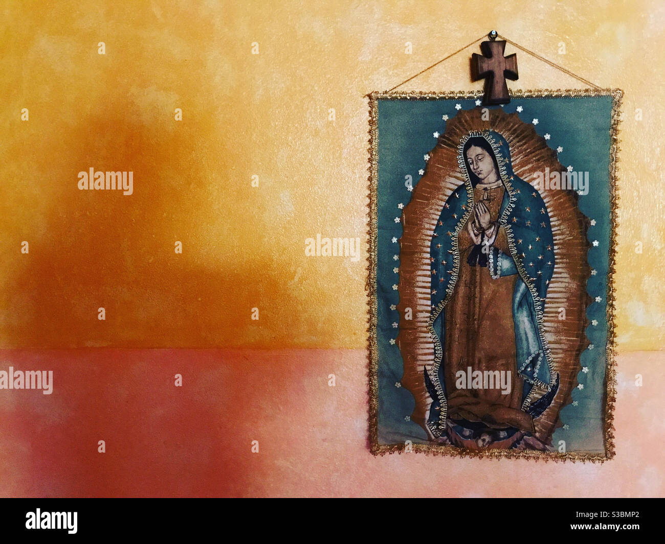 Ein mit einem christlichen Kreuz geschmücktes Banner der Muttergottes von Guadalupe in einem Haus in Peña de Bernal, Queretaro, Mexiko, wirft einen Schatten in eine orangefarbene Wand. Stockfoto