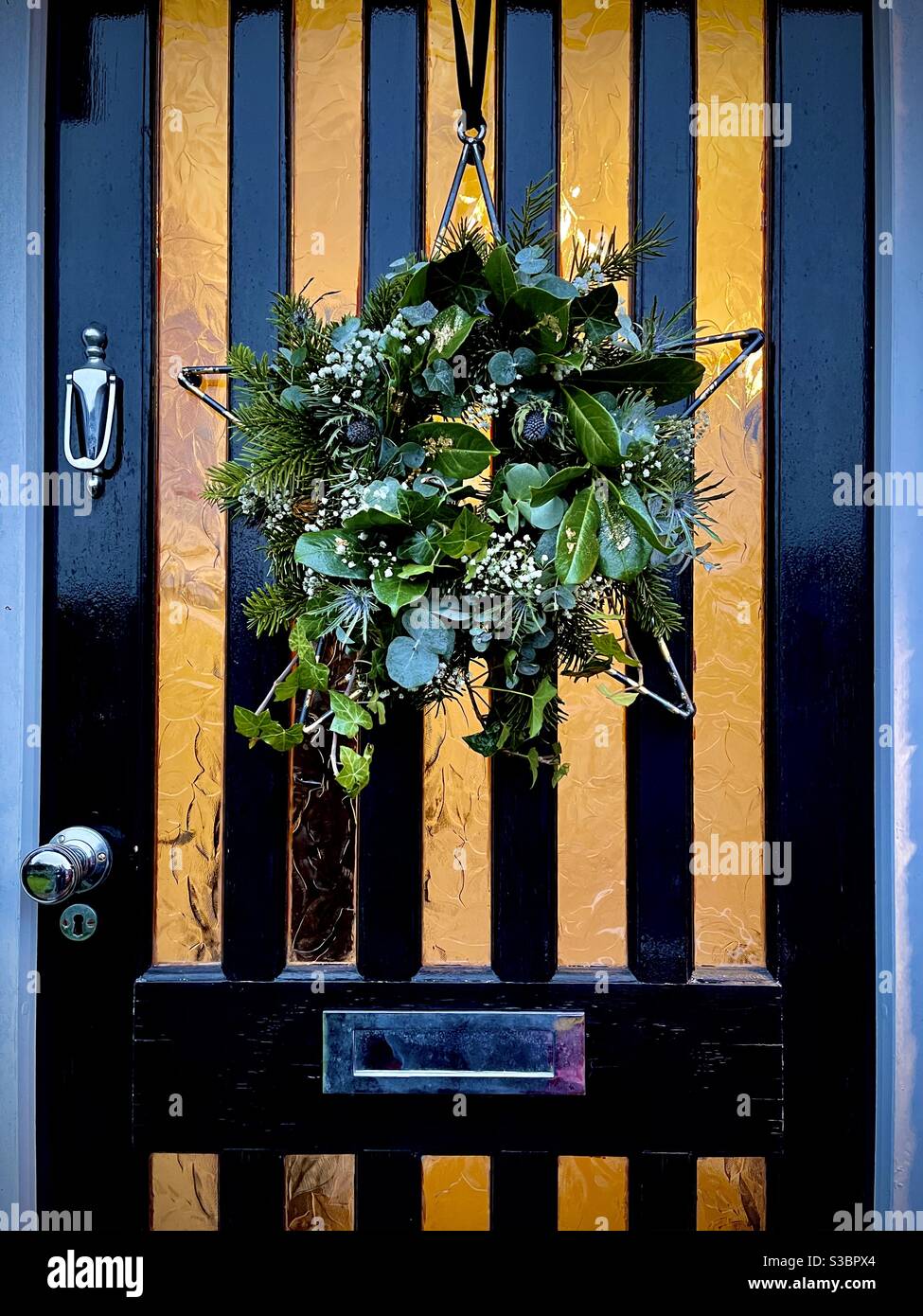 Weihnachtskranz mit Metallstern, der auf schwarz lackierter Vorderseite hängt Tür mit Licht im Haus Stockfoto