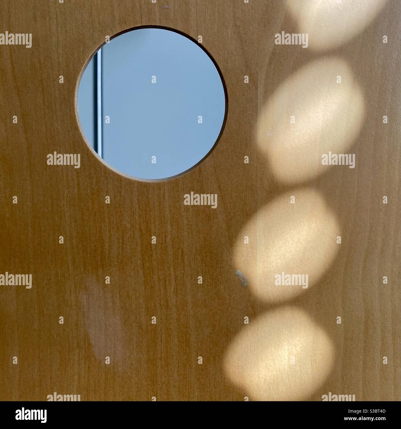 Lichtmuster, das durch Sonneneinstrahlung auf die Rückseite eines gebogenen Holzstücks mit einem Loch oben rechts und einem unscharf dahinter liegenden Objekt entsteht, was einen interessanten abstrakten Hintergrund erzeugt - Smartphone-aufgenommenes Stockfoto