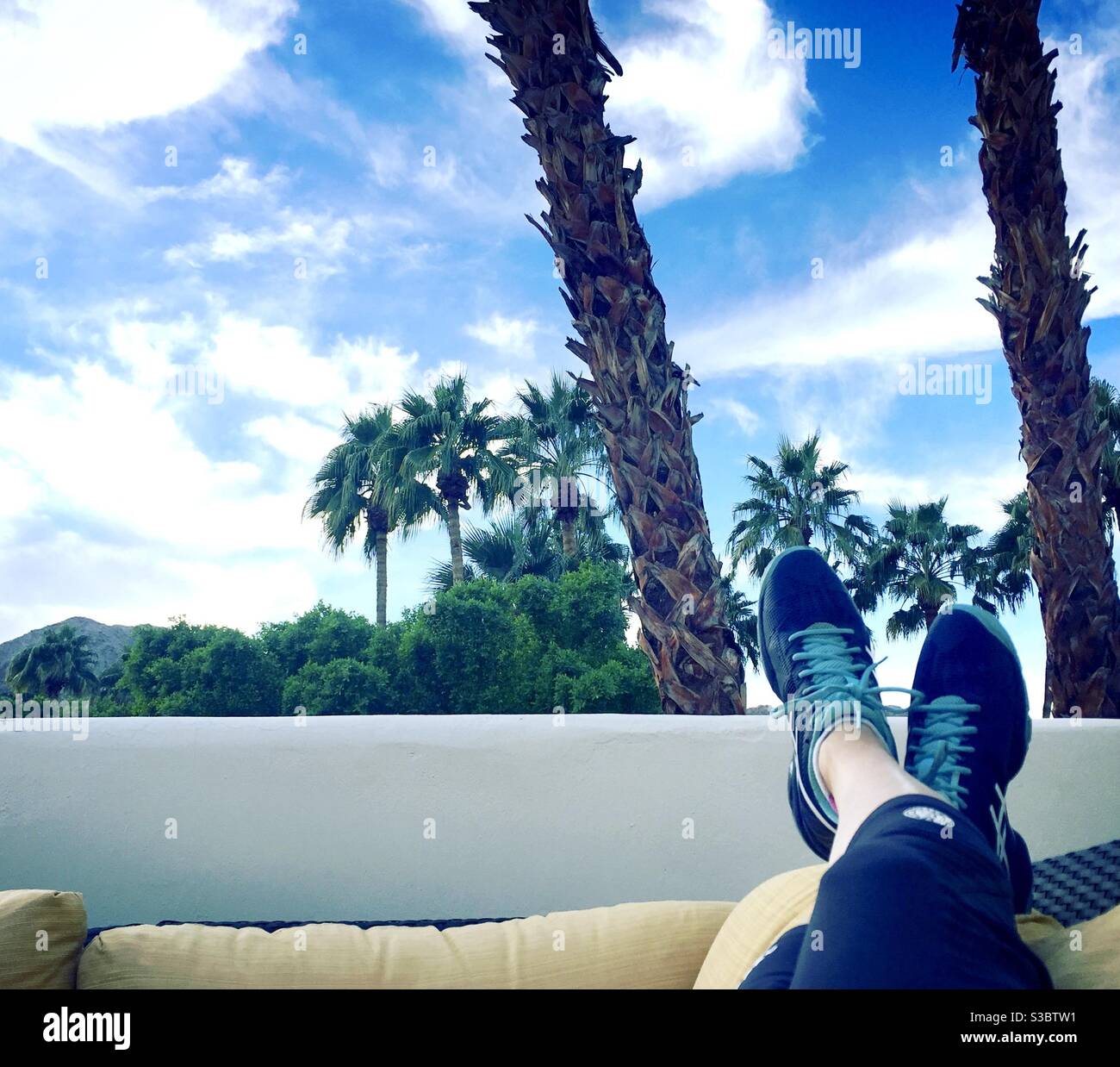 Entspannen in Palm Desert, Kalifornien. Beine und Füße tragen gekreuzte Tennisschuhe und ruhen auf gelbem Kissen mit Wüstenlandschaft im Hintergrund. Stockfoto