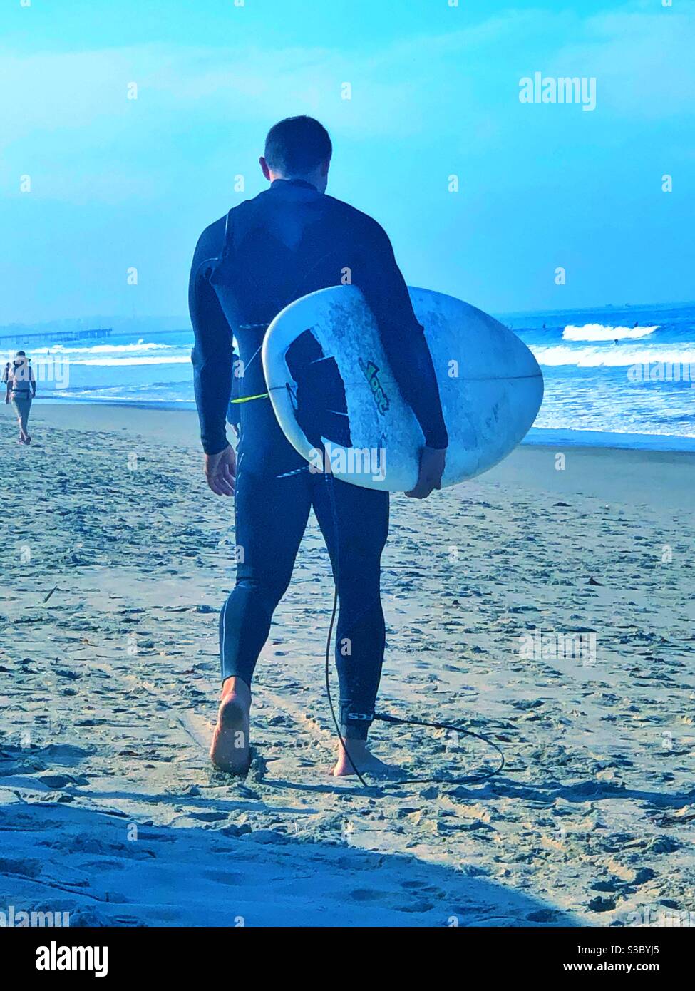 Kräftiger, muskulöser, dunkelhaariger junger Mann in einem Neoprenanzug, der ein Surfbrett am Strand in Santa Monica, CA, USA trägt und an einem kalten, klaren Tag in die Wellen des Pazifischen Ozeans geht. - Smartphone-aufgenommenes Stockfoto