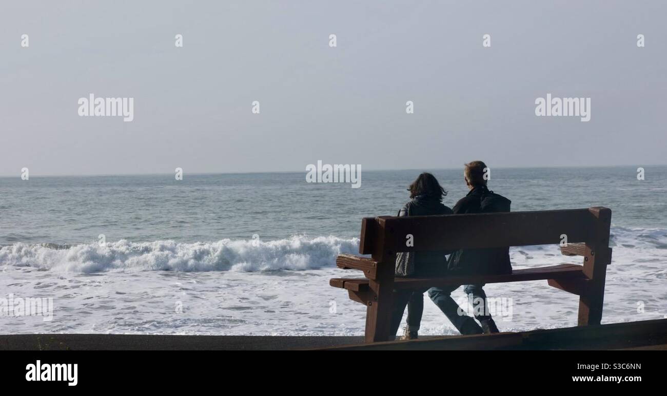 Ein Paar sitzt auf einer Bank neben einem Strand Stockfoto