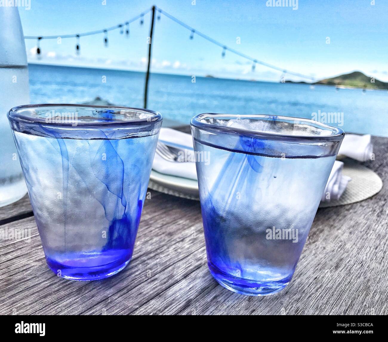 Zwei blaue Gläser voll Wasser auf einem Tisch mit Das Karibische Meer in Antigua im Hintergrund Stockfoto