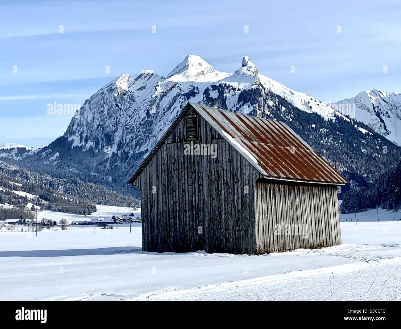 Alte Holzscheune mit rostigen Metalldach auf den Weiden rund um das Dorf Studen in der Schweiz im Winter mit viel Schnee. Detailansicht mit Bergen auf den Hintergrunsen. Stockfoto