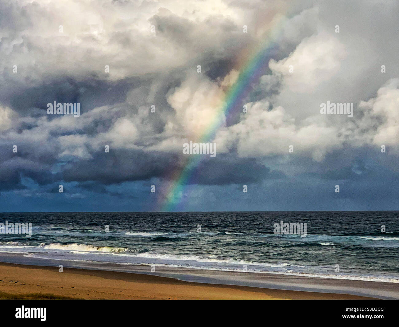 Schöner Regenbogen über dem Sandstrand, der sich vor einem grauen, stürmischen Himmel absticht, der ins Meer, Wellen, Natur und Australien führt - Smartphone-aufgenommenes Stockfoto