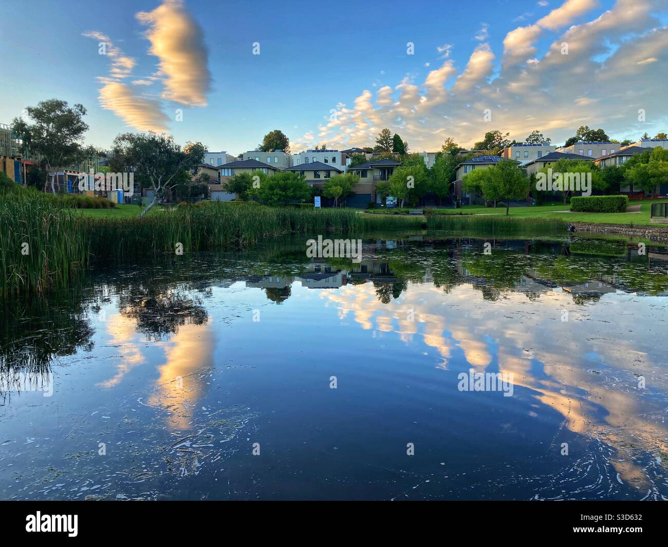 Wolken, die vom Morgenlicht in einem Teich reflektiert werden, umgeben von entfernten Häusern. Stockfoto