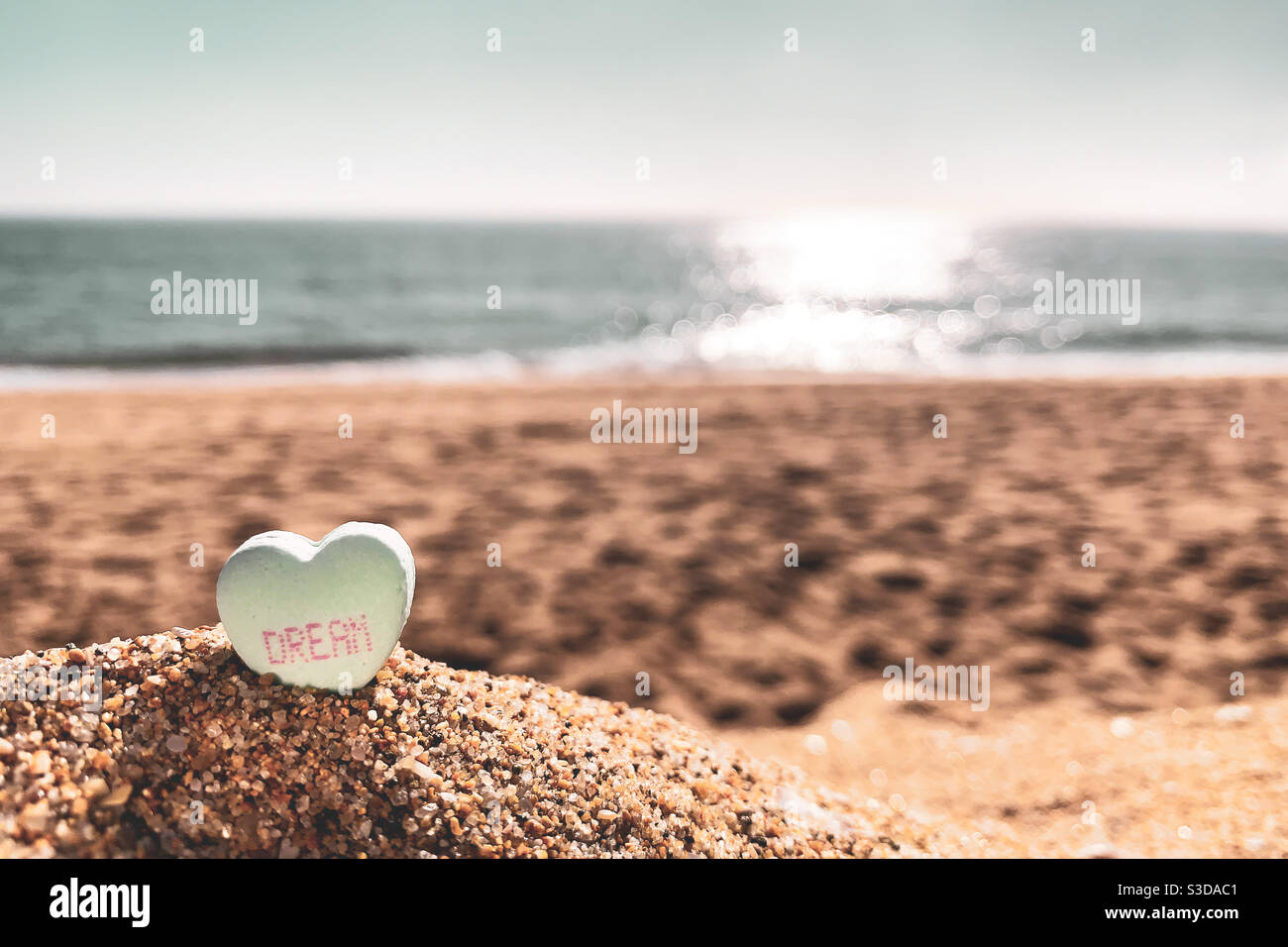 Bonbon Herz mit dem Wort "Traum" darauf, sitzen im Sand eines sonnigen Strandes mit dem Ozean verschwommen in der Ferne. Stockfoto