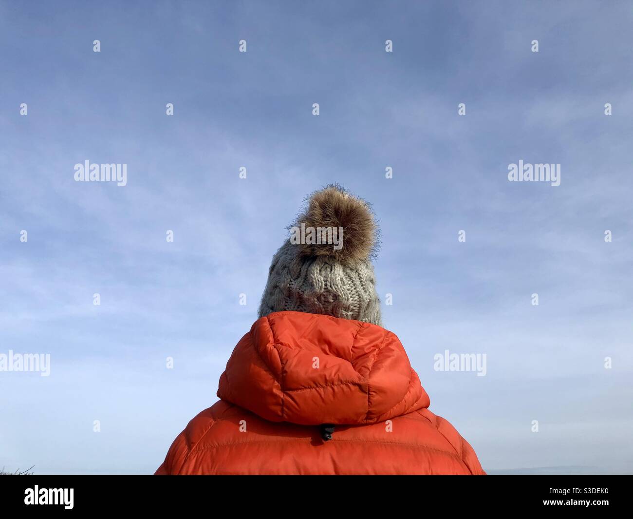 Rückansicht einer Frau im Freien, die einen warmen Winter trägt Mantel und gestrickter wolliger Bommel Hut mit blauem Himmel und Speicherplatz kopieren - Smartphone-aufgenommenes Stockfoto
