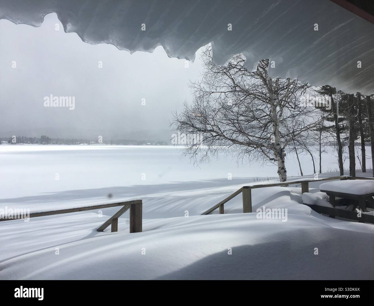 Eis gleitet vom Dach überhängende Terrasse und Picknick-Tisch unter Schnee begraben mit Blick auf Portage Lake im Norden von Maine, Aroostook County am 9. März 2018 nach einem Nor’easter. Stockfoto