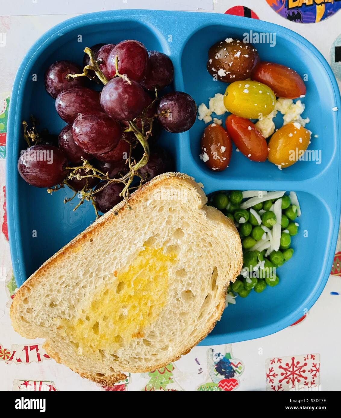 Ein einfaches, aber gesundes Mittagessen für ein Kind mit frischen roten Trauben, Kirschtomaten mit Feta-Käse, frischen Erbsen mit Mozzarella und einer Scheibe Toast mit Butter auf einem blauen Teller Stockfoto