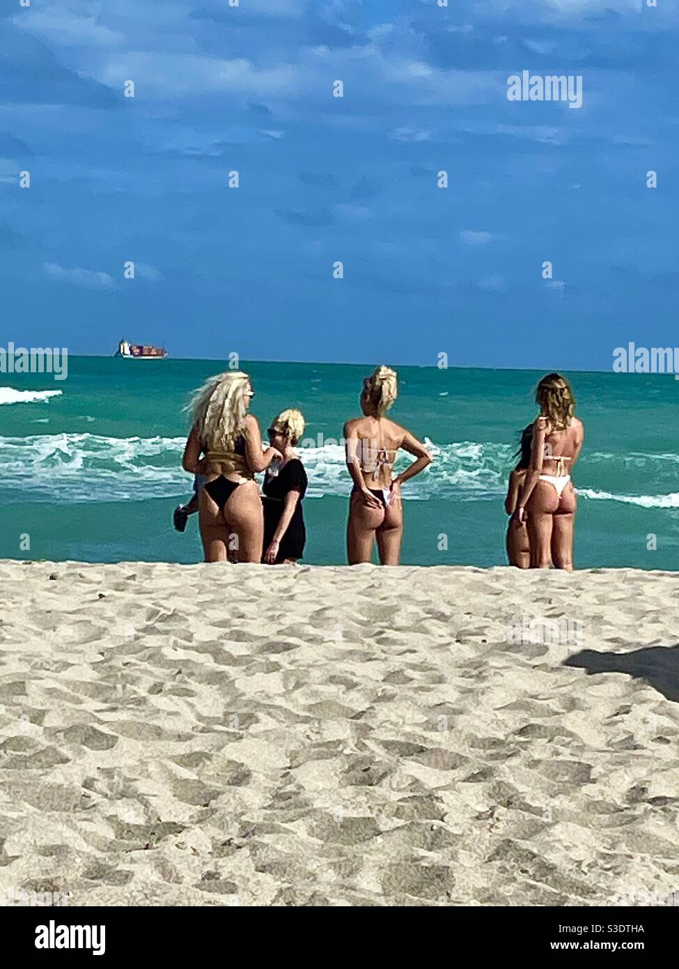 Frauen in Bikinis am Strand beobachten an einem klaren Tag im Februar ein Frachtschiff am Horizont des türkisfarbenen Meerwassers in Miami South Beach, Florida, USA - Smartphone-aufgenommenes Stockfoto