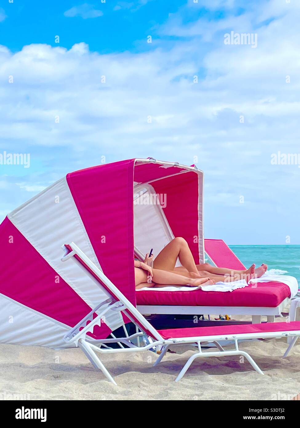 Gebräunte Beine ragen aus einem leuchtend rosa und weißen Cabana-Regenschirm im Beach Resort des SLS Hotels in Miami South Beach, Florida, USA Stockfoto