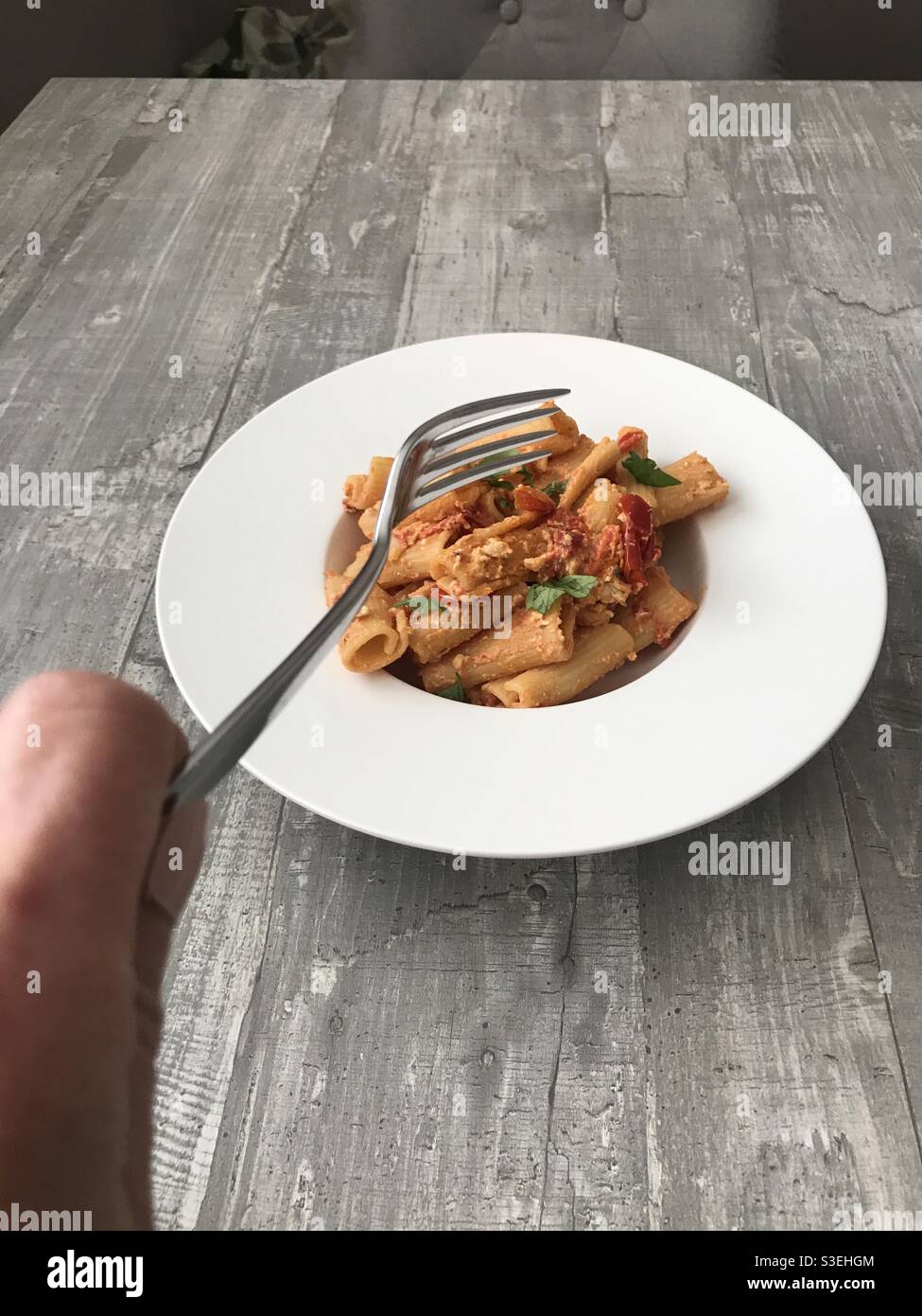 Frau, die gerade dabei ist, eine Gabel in eine Schüssel mit frisch zubereiteten Rigatoni mit Feta, Tomaten und Basilikumsoße zu legen - Smartphone-aufgenommenes Stockfoto