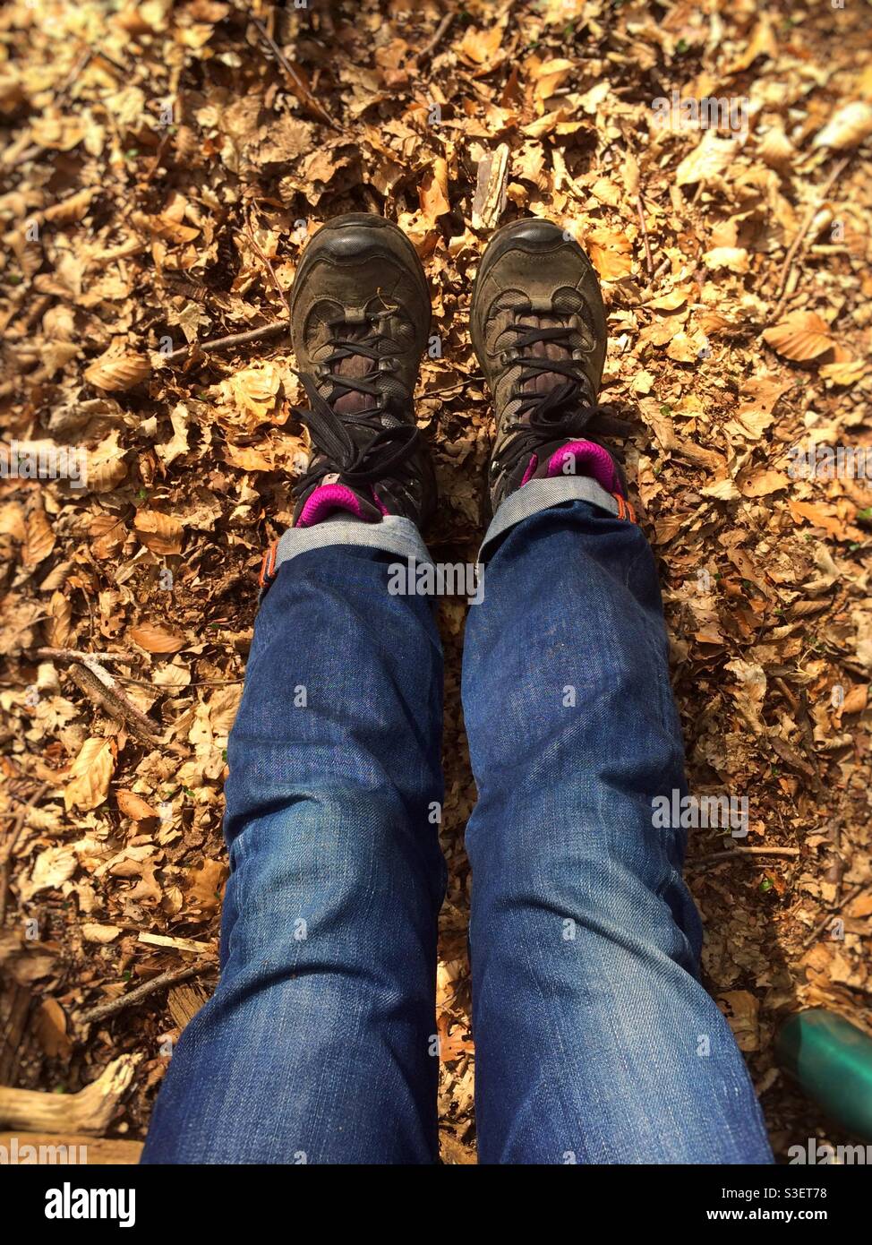 Die Beine einer Frau sind vor ihr ausgestreckt, während sie auf dem mit Blättern übersäten Waldboden sitzt. Sie trägt blaue Jeans und Wanderschuhe. Stockfoto