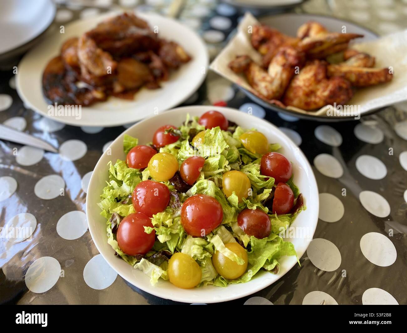 Salat mit roten und grünen Minitomaten auf Salat, mit Hähnchenflügeln auf einem schwarz-weiß gepunkteten Plastiktischtuch Stockfoto