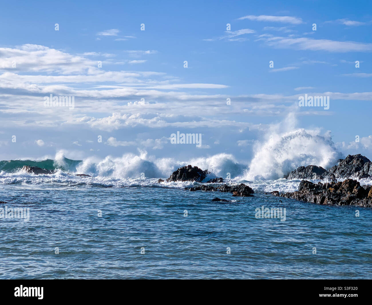 Schöner blauer Himmel am Strand, ein Bogen aus weiß schaumigen Wasser von Wellen, die über Felsen in der Nähe der Küste krachen. Meeresspray fliegt. Sawtell Australien - Smartphone-aufgenommenes Stockfoto