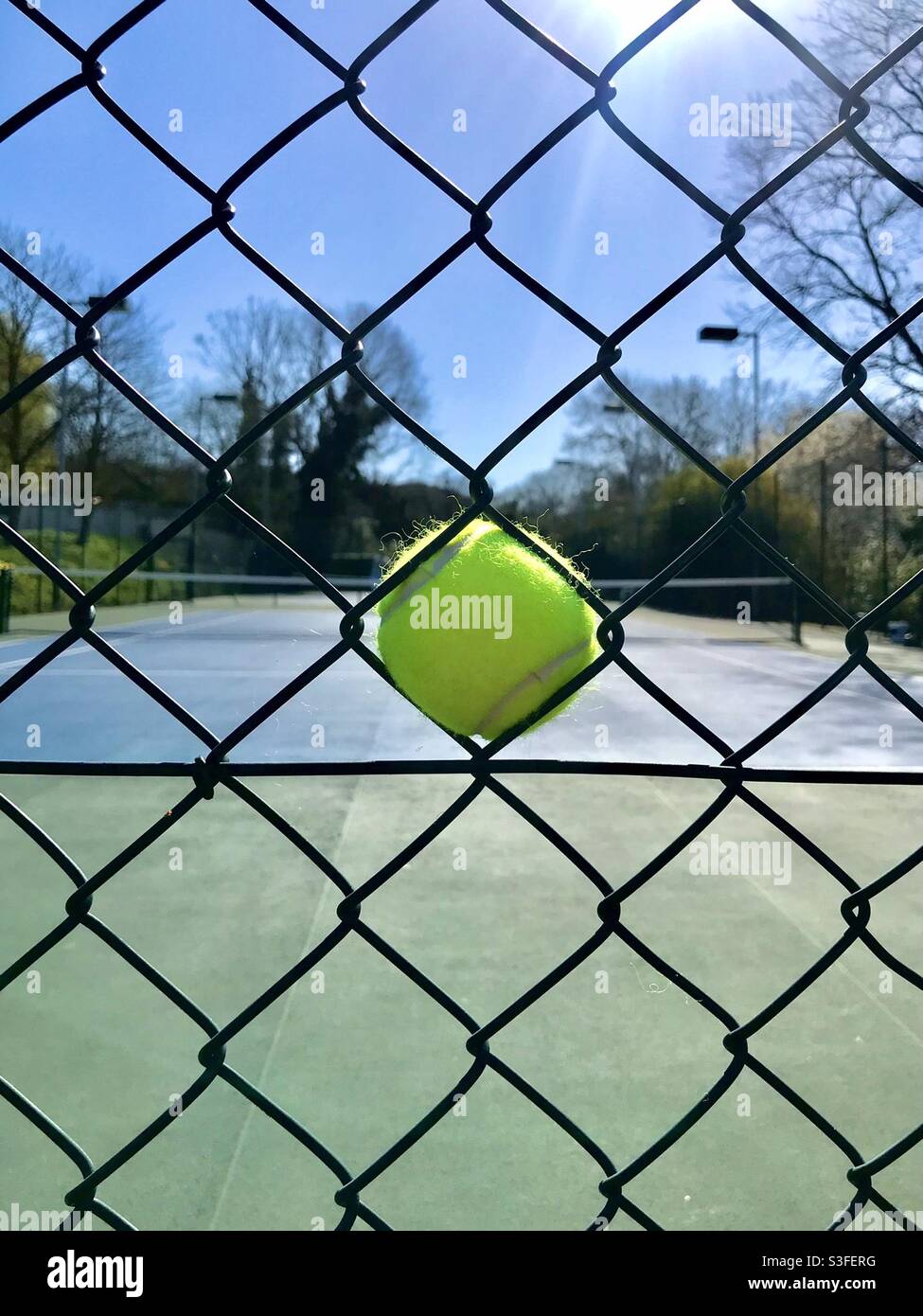 Blick auf einen Tennisplatz durch einen Zaun, mit einem Tennisball in der Mitte, an einem wunderschönen sonnigen Tag - Smartphone-aufgenommenes Stockfoto