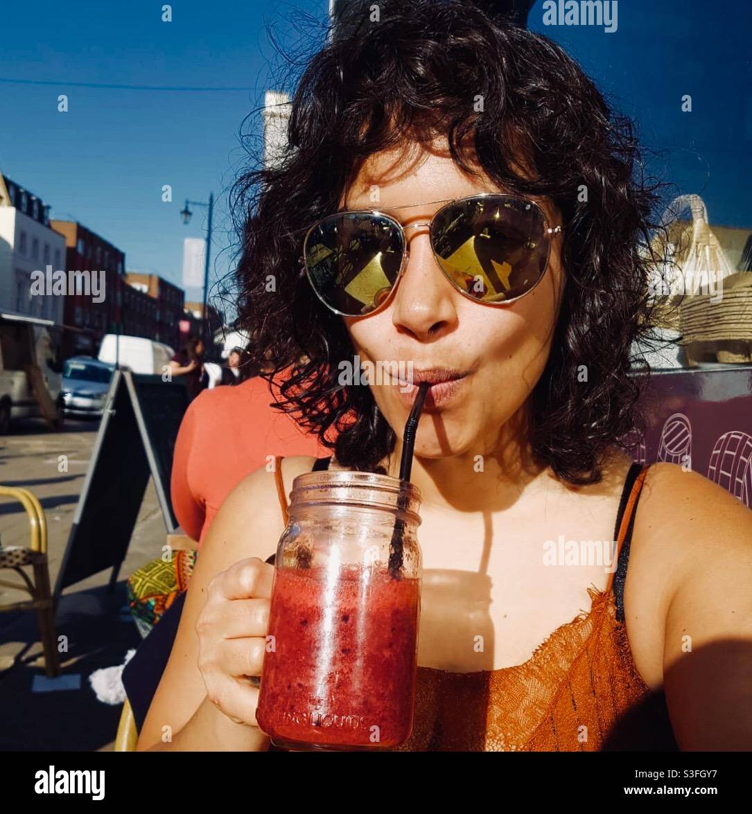 Lockig haarige Mädchen große Sonnenbrille trinken roten Cocktail Smoothie in einem Glas in der Sonne. Stockfoto