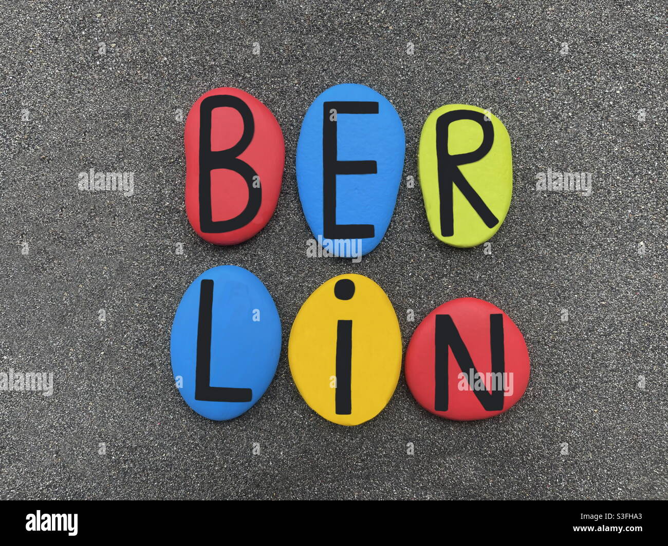 Berlin, kreatives Logo mit handgefertigten farbigen Steinbuchstaben über natürlichem schwarzen Vulkansand Stockfoto