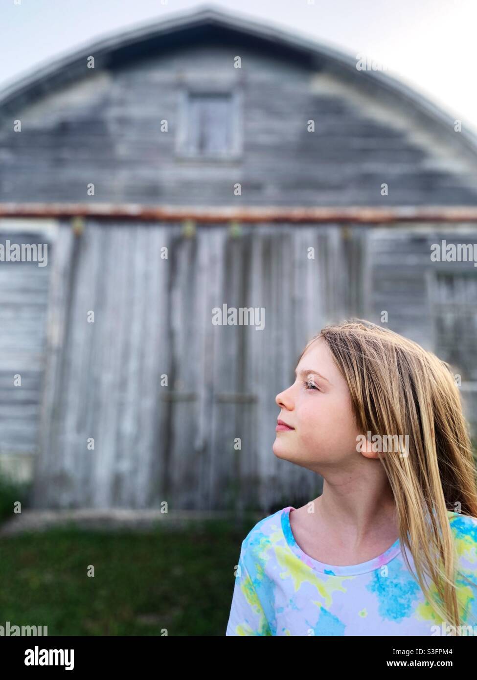 Ein junges blondes Kind, das in der Ferne vor einer alten Scheune steht Stockfoto