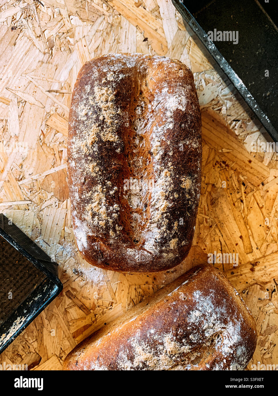 Zwei Getreidebrote und ihre Backformen auf Spanplatten-Hintergrund - Smartphone-aufgenommenes Stockfoto