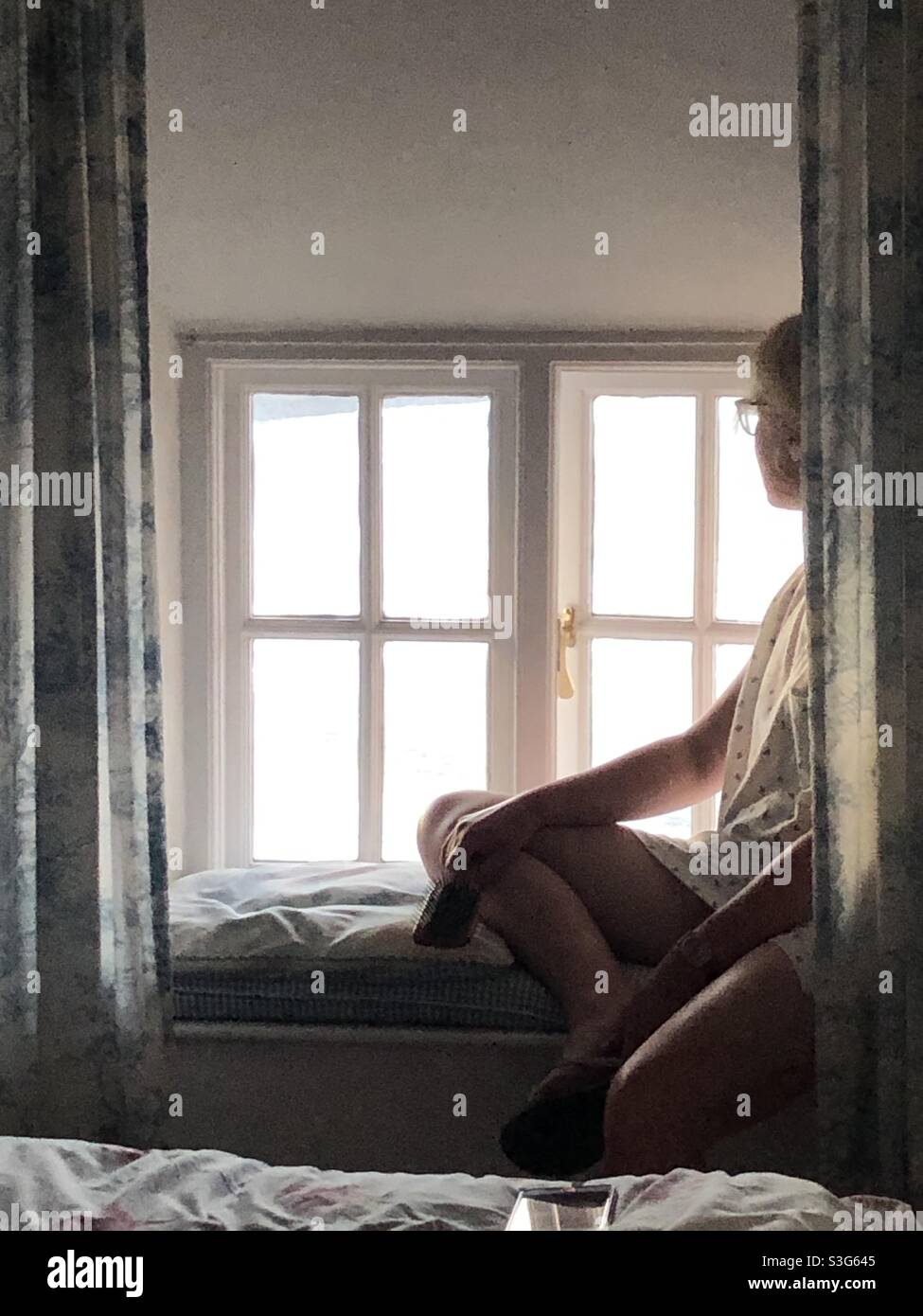 Eine einsame Frau, die an einem Fensterplatz in ihrem Schlafzimmer sitzt und aus dem Fenster schaut, und ein Konzept für die psychische Gesundheit Stockfoto