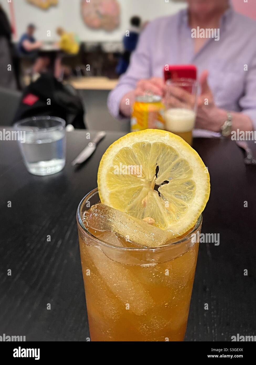 Hohes Glas erfrischenden Eistees mit einer großen Zitronenscheibe auf einem Tisch vor einem Mann, der mit seinem Handy spielt, USA - Smartphone-aufgenommenes Stockfoto