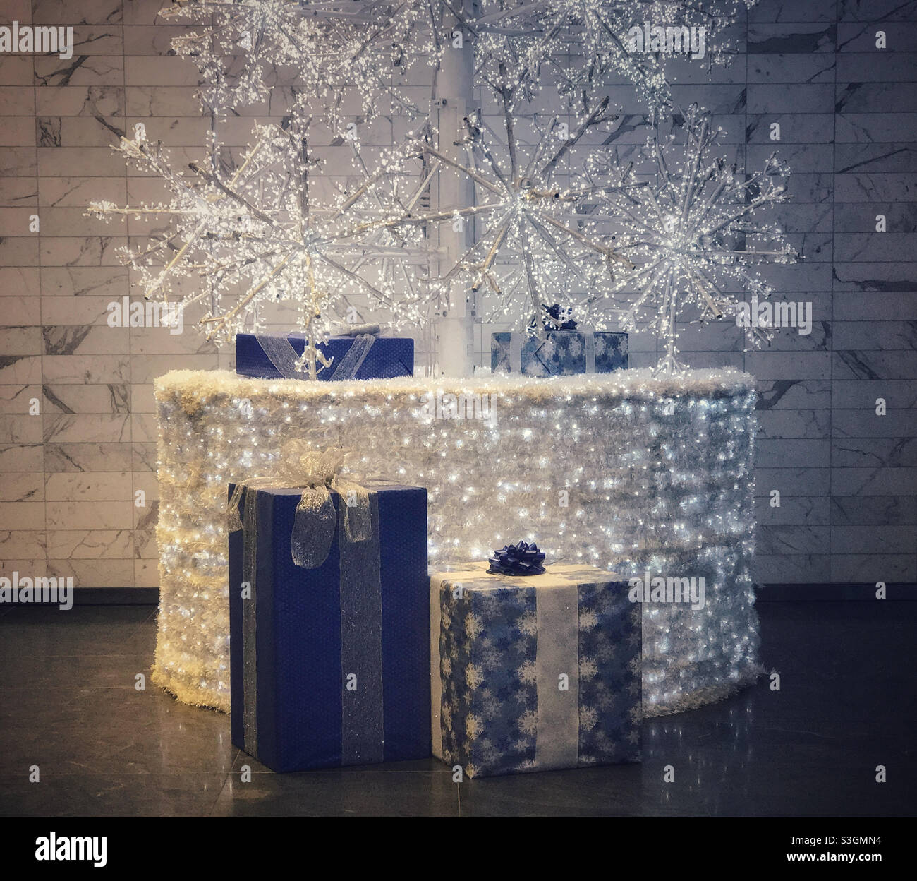 Geschenke in blauem Geschenkpapier verpackt, am Fuß eines beleuchteten weißen künstlichen Weihnachtsbaums. - Smartphone-aufgenommenes Stockfoto