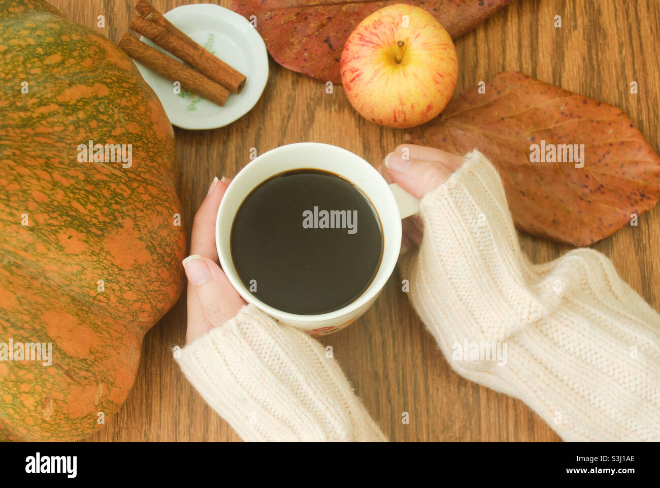 Eine Person, die eine Tasse Kaffee, einen Apfel, Zimtstangen, Kürbis und braune Blätter auf einem Holzschreibtisch hält Stockfoto