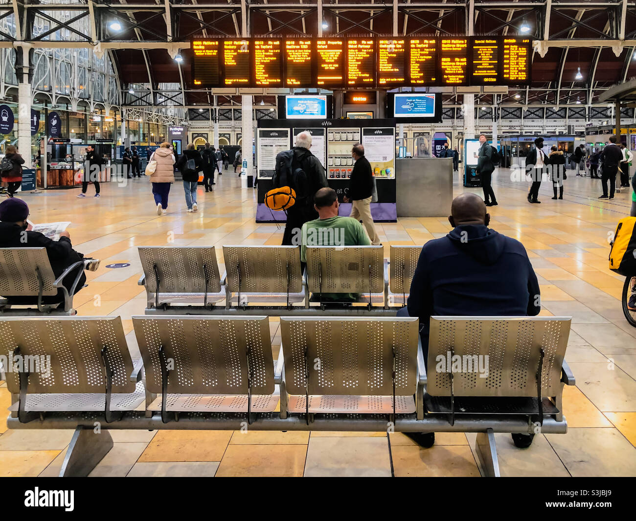 Die Leute sitzen auf Bänken und beobachten die Abfahrtstafeln in der Bahnhofskonbahn am Bahnhof Paddington in London, Großbritannien. Stockfoto