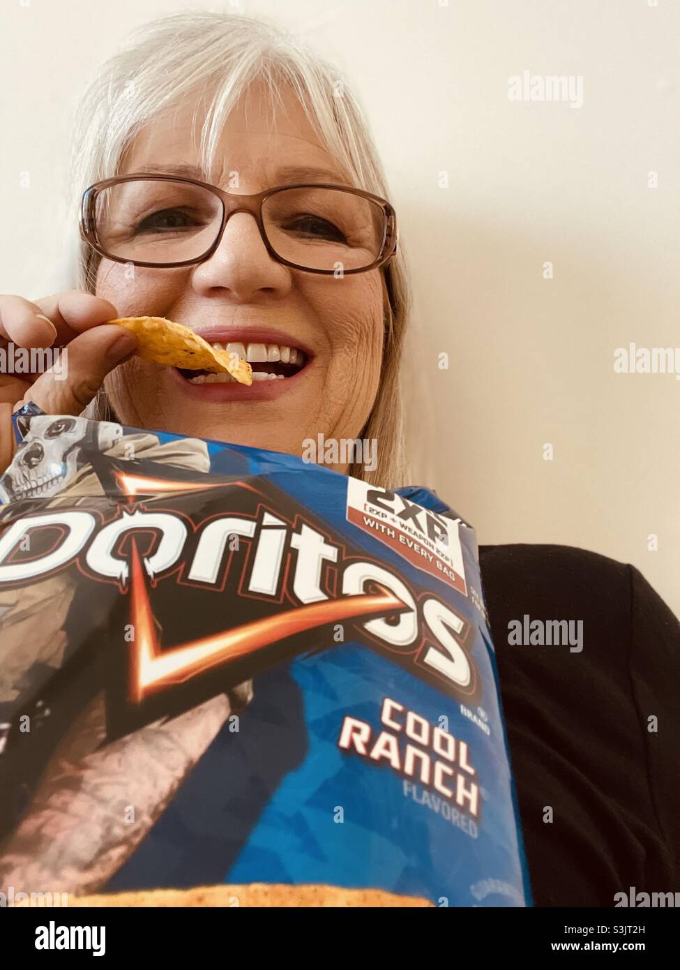 Hellhaarige Frau in Gläsern lächelt, als sie Doritos coole Ranch-Chips isst Stockfoto