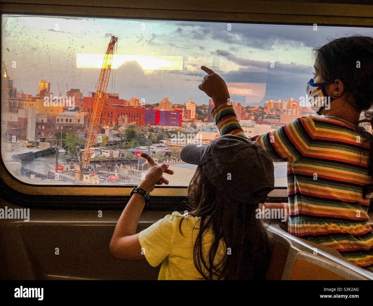 Zwei junge Mädchen, die bei Sonnenuntergang durch das Fenster des F-Zuges zum Gowanus-Kanal und in die Innenstadt von Brooklyn blicken Stockfoto