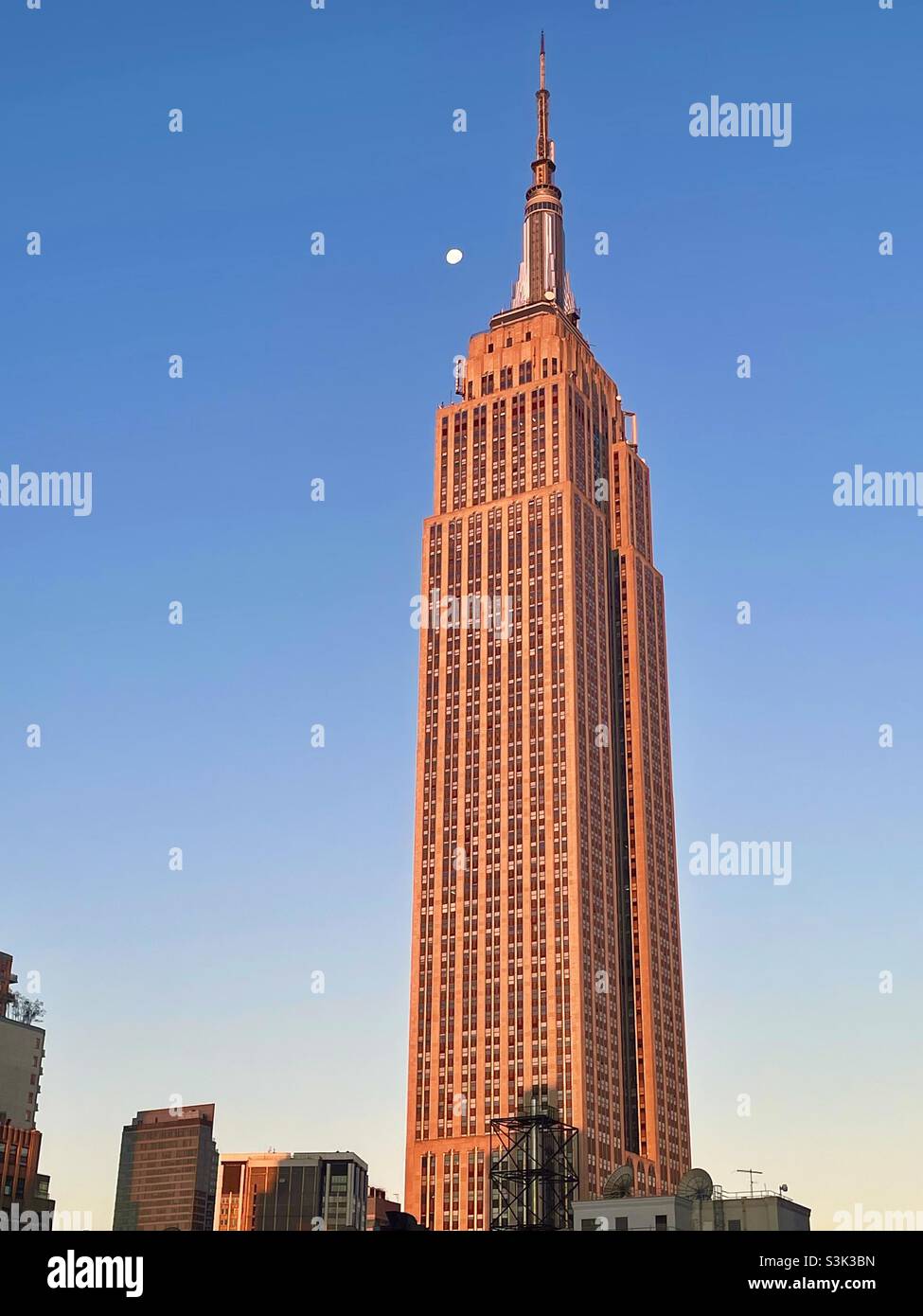 The Moon & New York City: Das Licht am frühen Morgen wirft ein rosa Leuchten auf das Empire State Building. Stockfoto