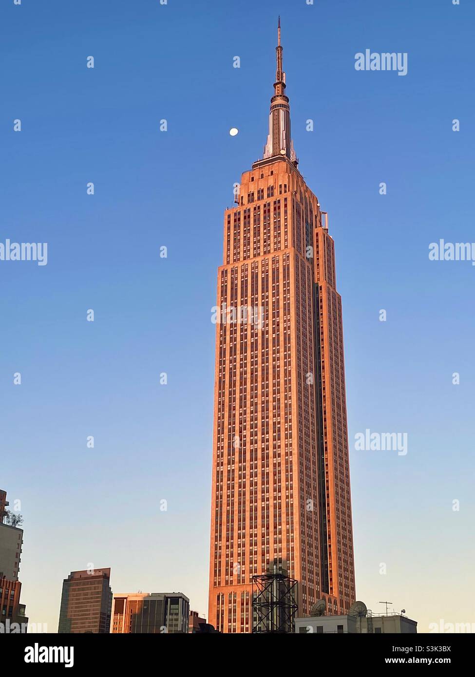 Blick auf das Empire State Building am frühen Morgen, wobei das Sonnenlicht einen rosa Glanz auf den Mond wirft, der sich dem Gebäude nähert Stockfoto