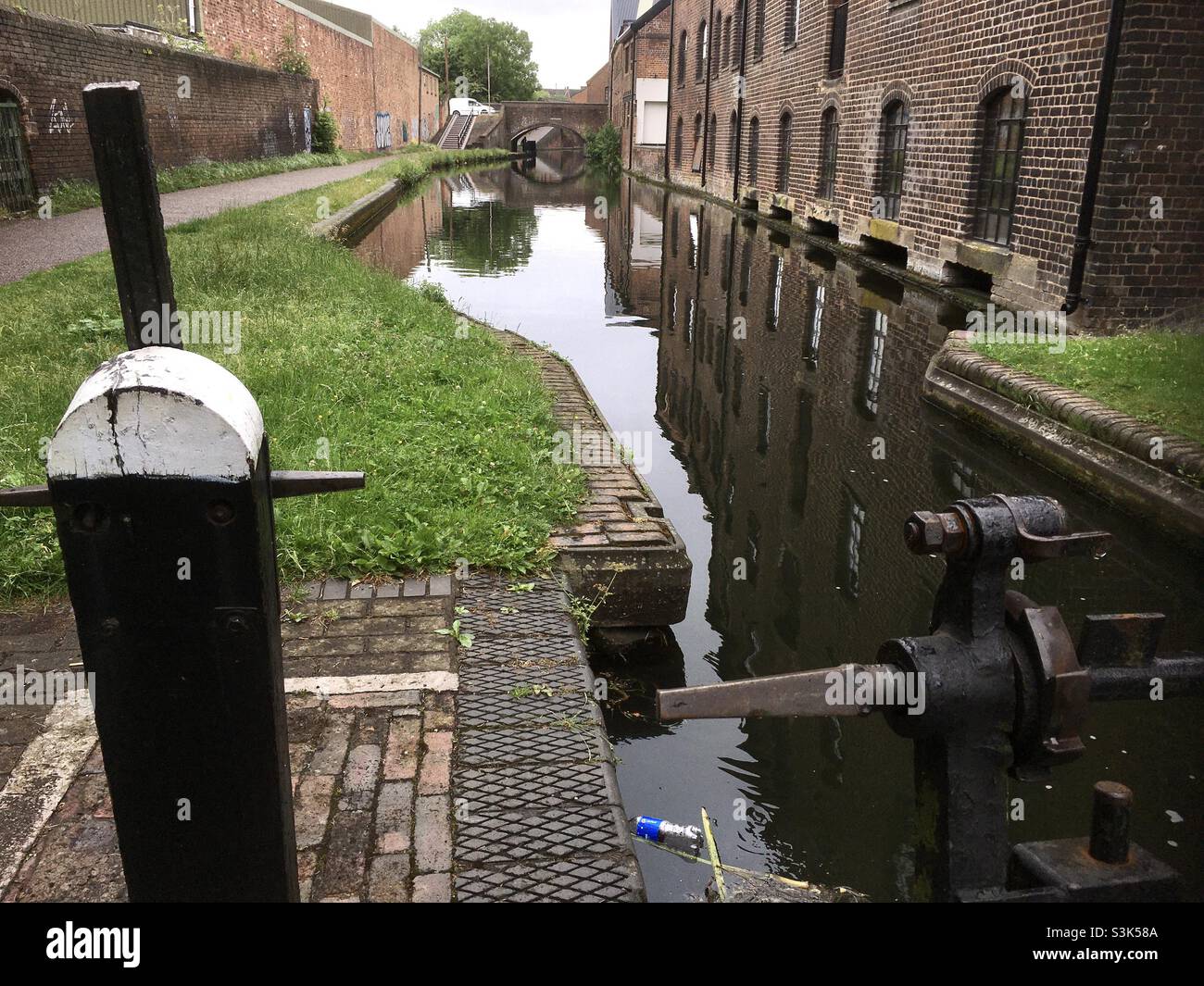 Blick entlang des Stourbridge-Kanals, der Gebäudereflexionen von Pfad und Brücke in der Ferne zeigt, mit dem Schleusenmechanismus im Vordergrund. - Smartphone-aufgenommenes Stockfoto