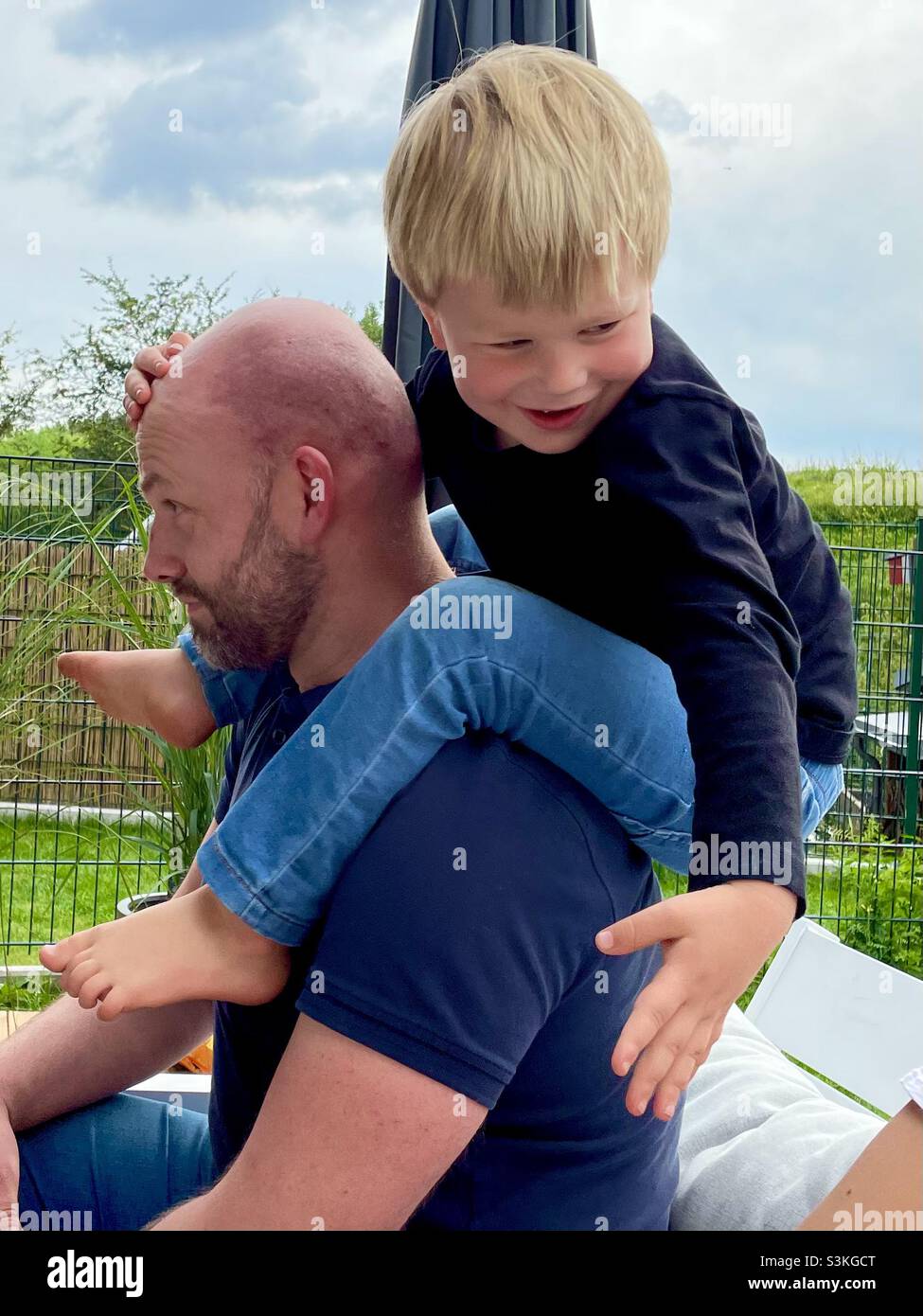 Lachendes Kleinkind, das auf den Schultern eines kahlen Mannes sitzt Stockfoto