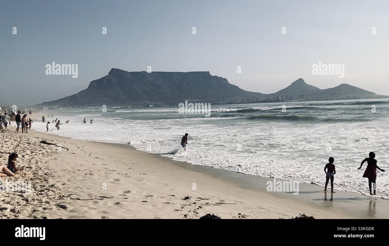 Tafelberg & Signalhügel bei Flut vom Milnerton Beach, Kapstadt. Badegäste genießen die Brandung. Stockfoto