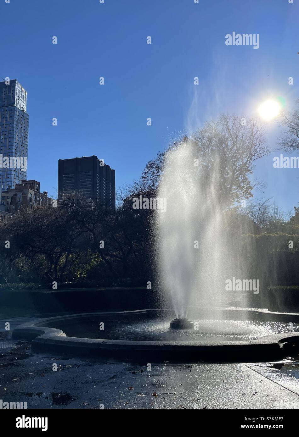 Der Wasserbrunnen wird von der Sonne gegen die Silhouetten der Wolkenkratzer in New York City beleuchtet - Smartphone-aufgenommenes Stockfoto