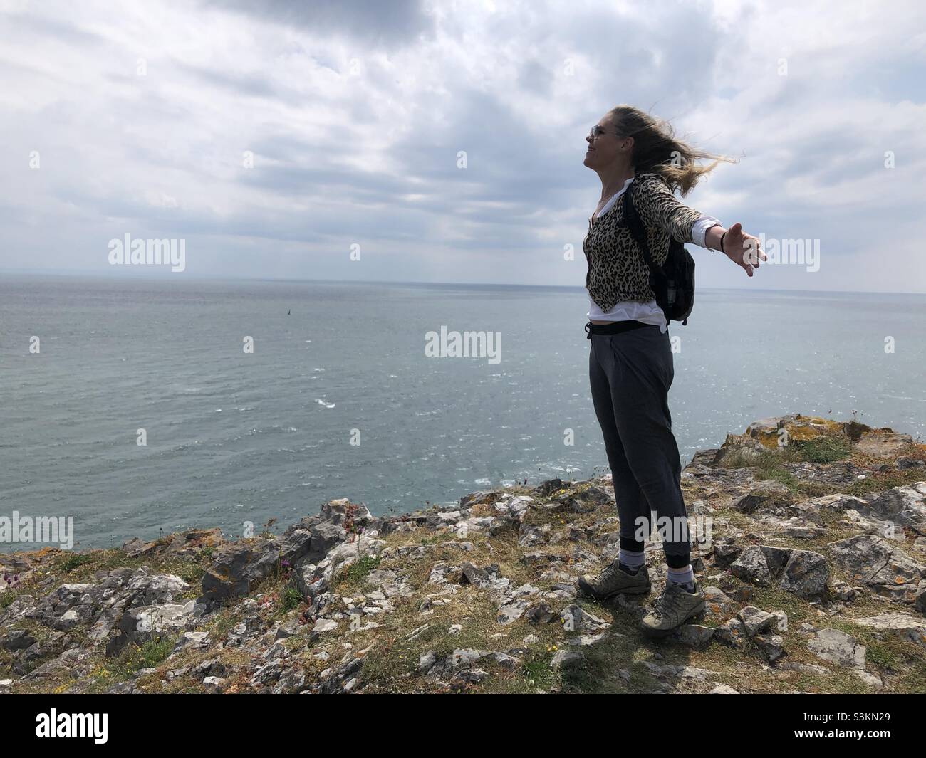 Auch in Südwales steht eine Frau mit weiten Armen vor starkem Wind auf einer Klippe - Smartphone-aufgenommenes Stockfoto