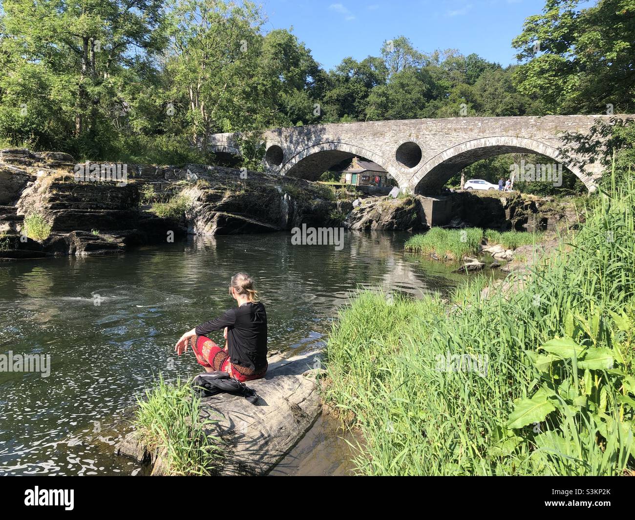 An einem sonnigen Tag in Wales sitzt eine Frau am Ufer eines Flusses in der Nähe der Brücke bei Salmons LEAP - Smartphone-aufgenommenes Stockfoto