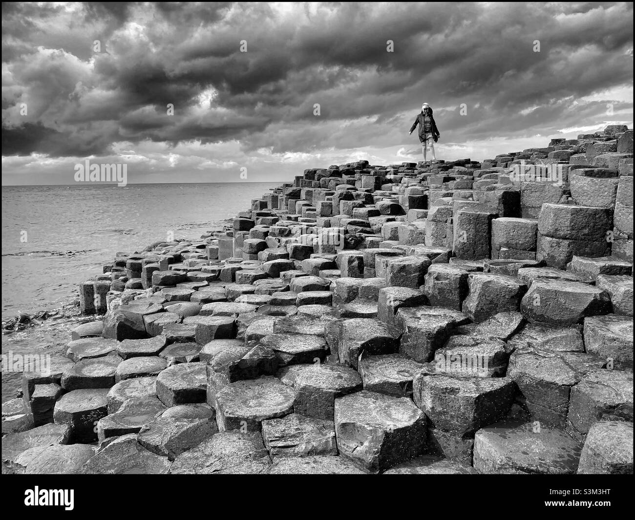 Ein einsame Wanderer geht über die vulkanischen Basaltsäulen, die das Gebiet Giant’s Causeway in Nordirland bilden. Ein sich näherender Sturm braut am Himmel. Foto ©️ COLIN HOSKINS. - Smartphone-aufgenommenes Stockfoto