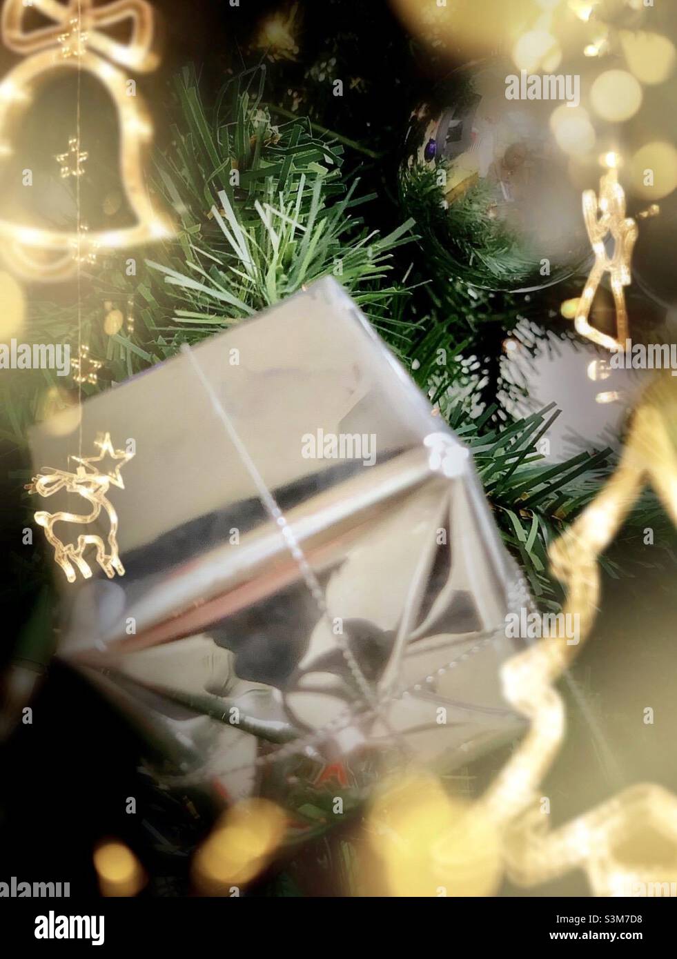 Auf einem Weihnachtsbaum ist eine silberne und glänzende Geschenkbox mit den gelblichen Lichtzeichnungen der Festschmuck um ihn herum ausgestellt. Stockfoto