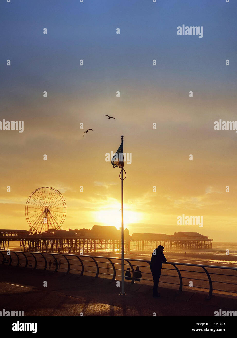 Figur in Silhouette, die an einer Barriere angelehnt ist und die Nachmittagssonne hinter dem Central Pier in Blackpool beobachtet Stockfoto