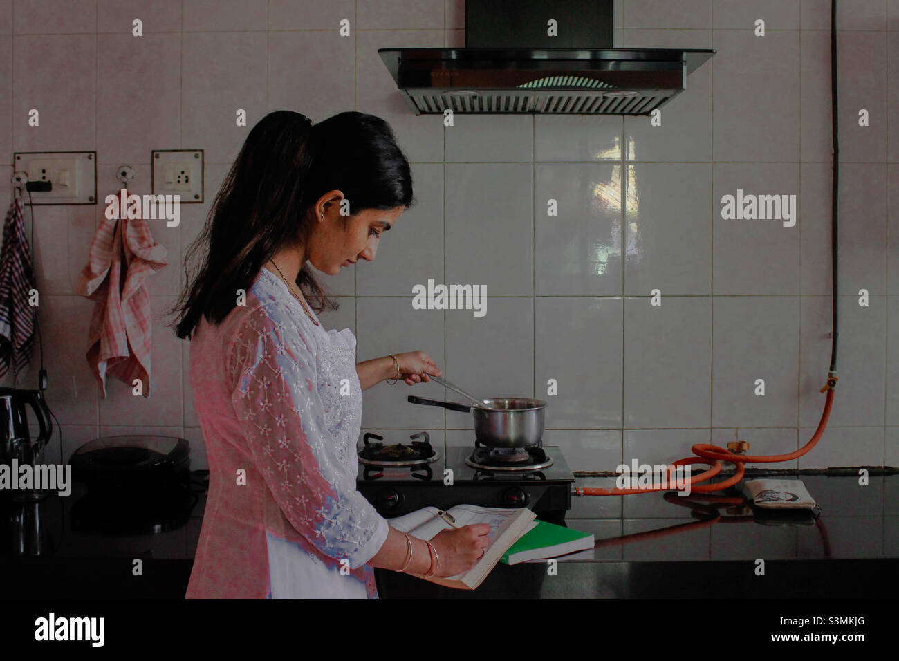 Dieses Bild zeigt eine südasiatische Frau, die studiert, während sie Hausarbeit unternimmt. Eine gelebte Erfahrung vieler ist, gleichzeitig studieren zu müssen, während sie mit Haushaltsverantwortung fortfährt Stockfoto