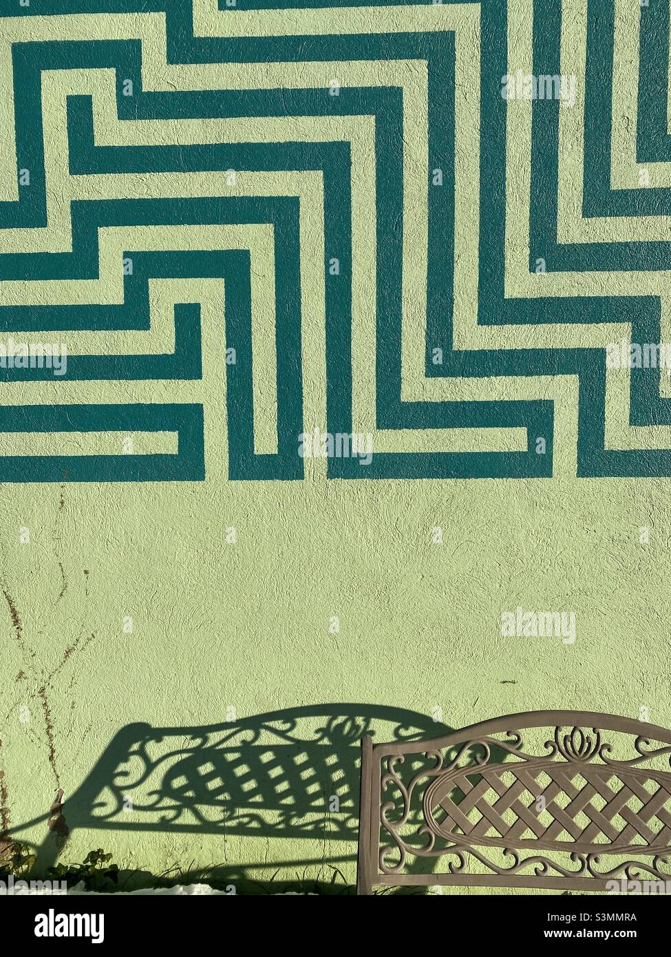 Ein grünes Labyrinth ist an der Seite eines Gebäudes gemalt, und eine Bank wirft einen ähnlichen Schatten auf die Wand darunter. - Smartphone-aufgenommenes Stockfoto