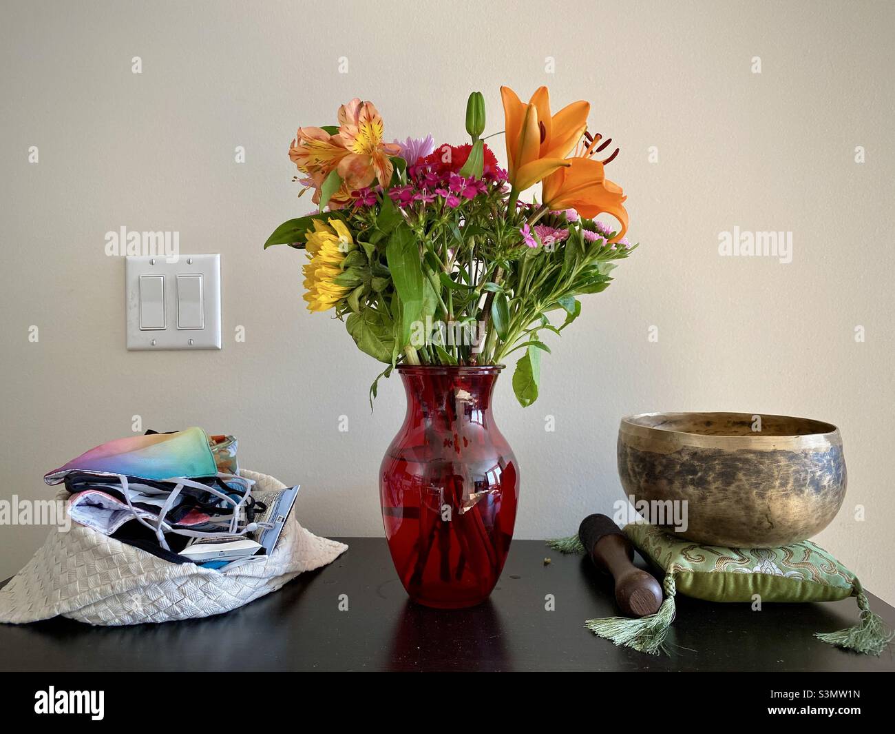 Blumenstrauß in roter Glasvase, auf dem Tisch mit Tüte kovidierter Masken und singender Messingschale auf beiden Seiten Stockfoto