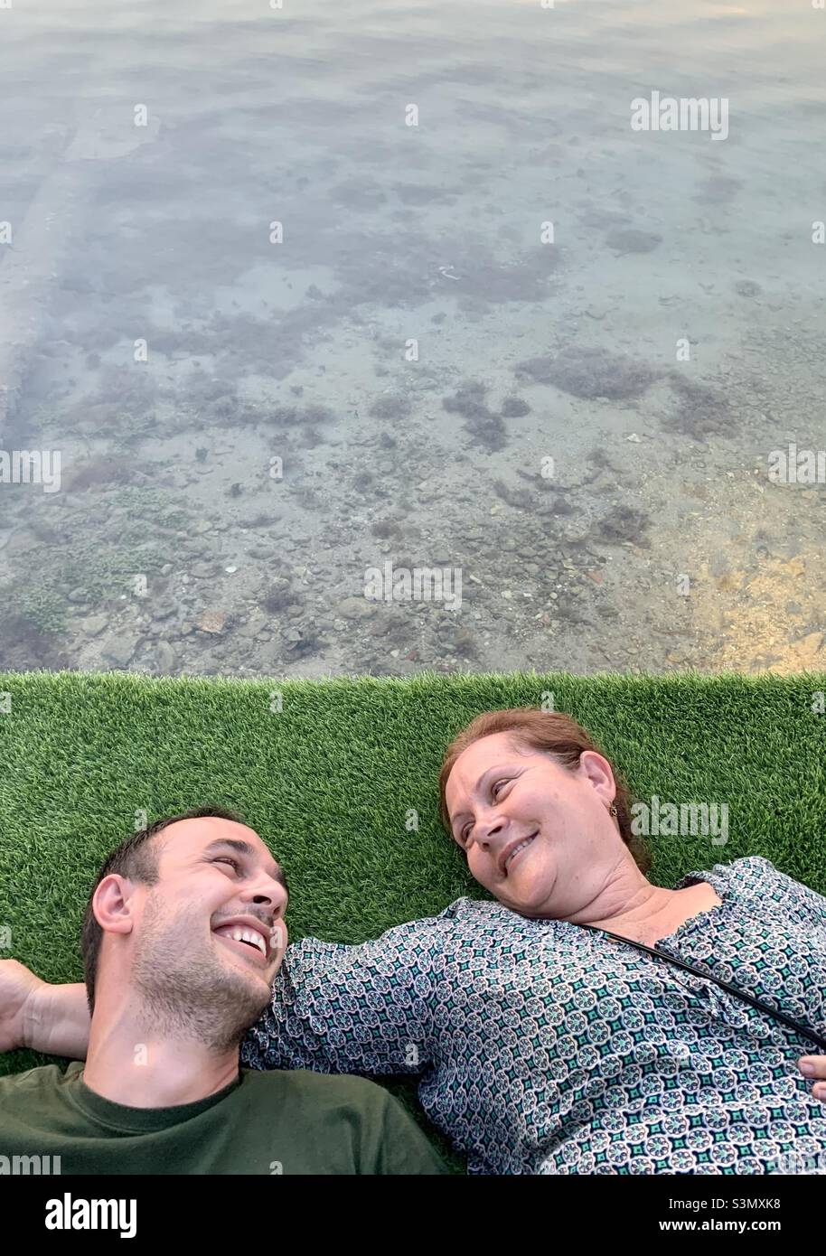 Mutter und Sohn lächeln einander an, während sie entspannt auf einem Grasfeld neben dem Meer liegen. Havanna, Kuba - Smartphone-aufgenommenes Stockfoto